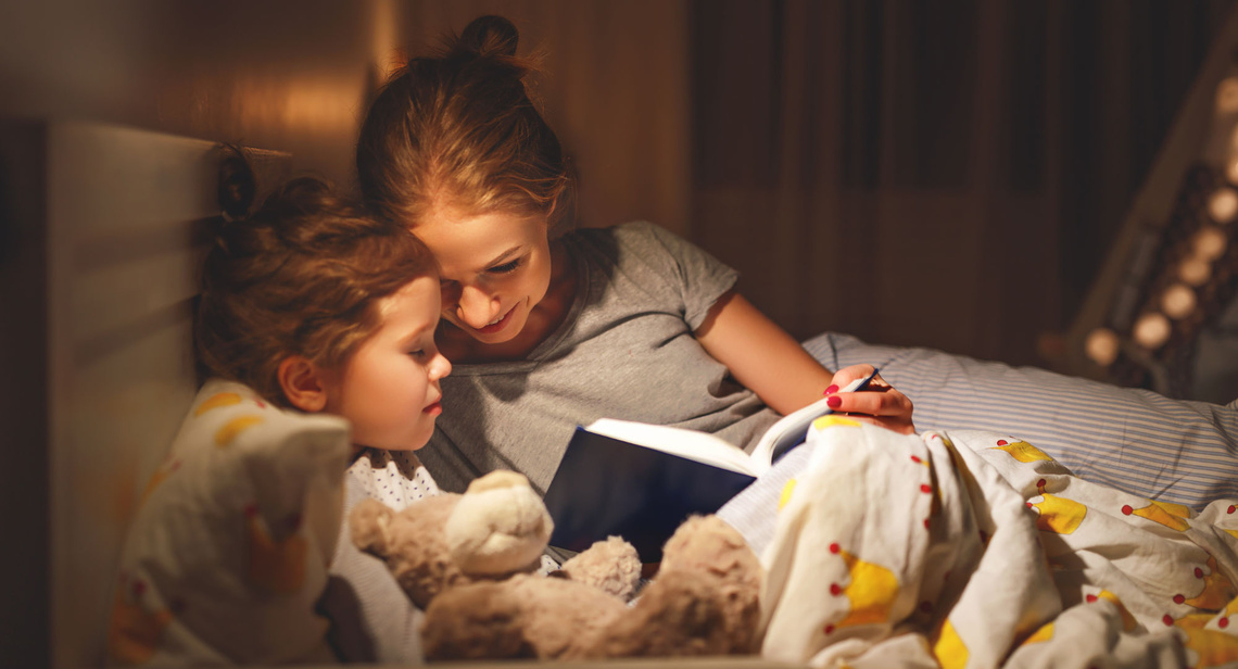 Mutter und Kind lesen bei Lampenlicht im Bett.