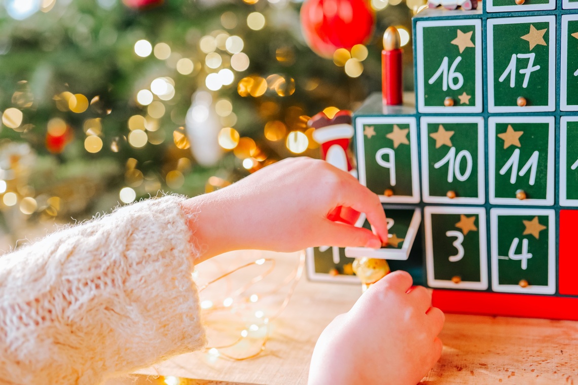 Eine Kinderhand öffnet ein Türchen an einem weihnachtlichen Adventskalender mit grün-weißen Zahlen und goldenen Sternen, im Hintergrund ein festlich geschmückter Weihnachtsbaum mit Lichtern.